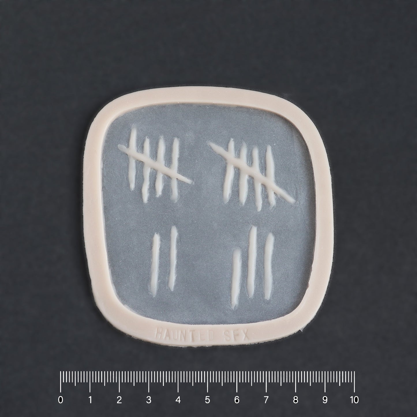 Tally Marks - Silicone makeup prosthetic in vanilla shade on a black surface with a ruler