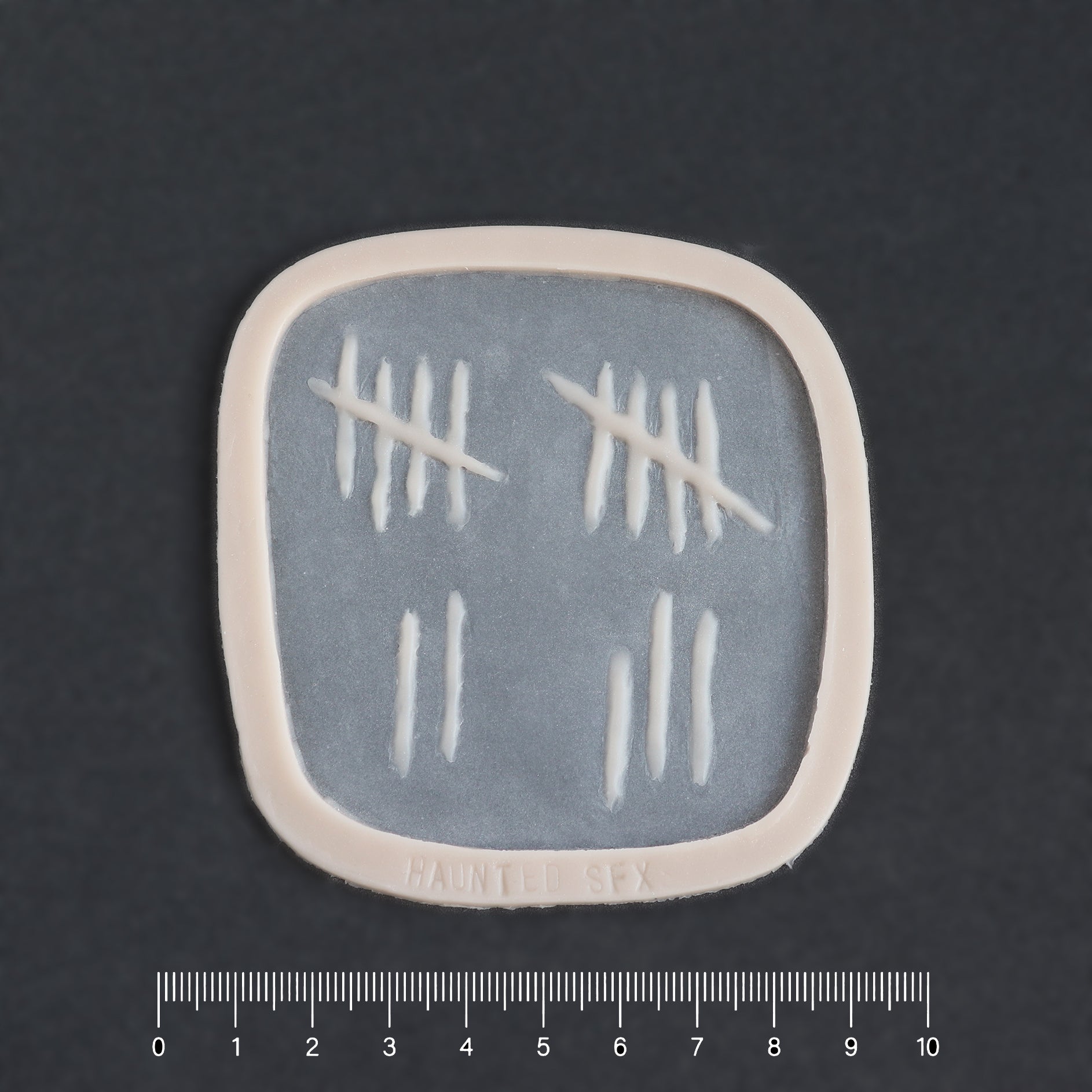 Tally Marks - Silicone makeup prosthetic in vanilla shade on a black surface with a ruler
