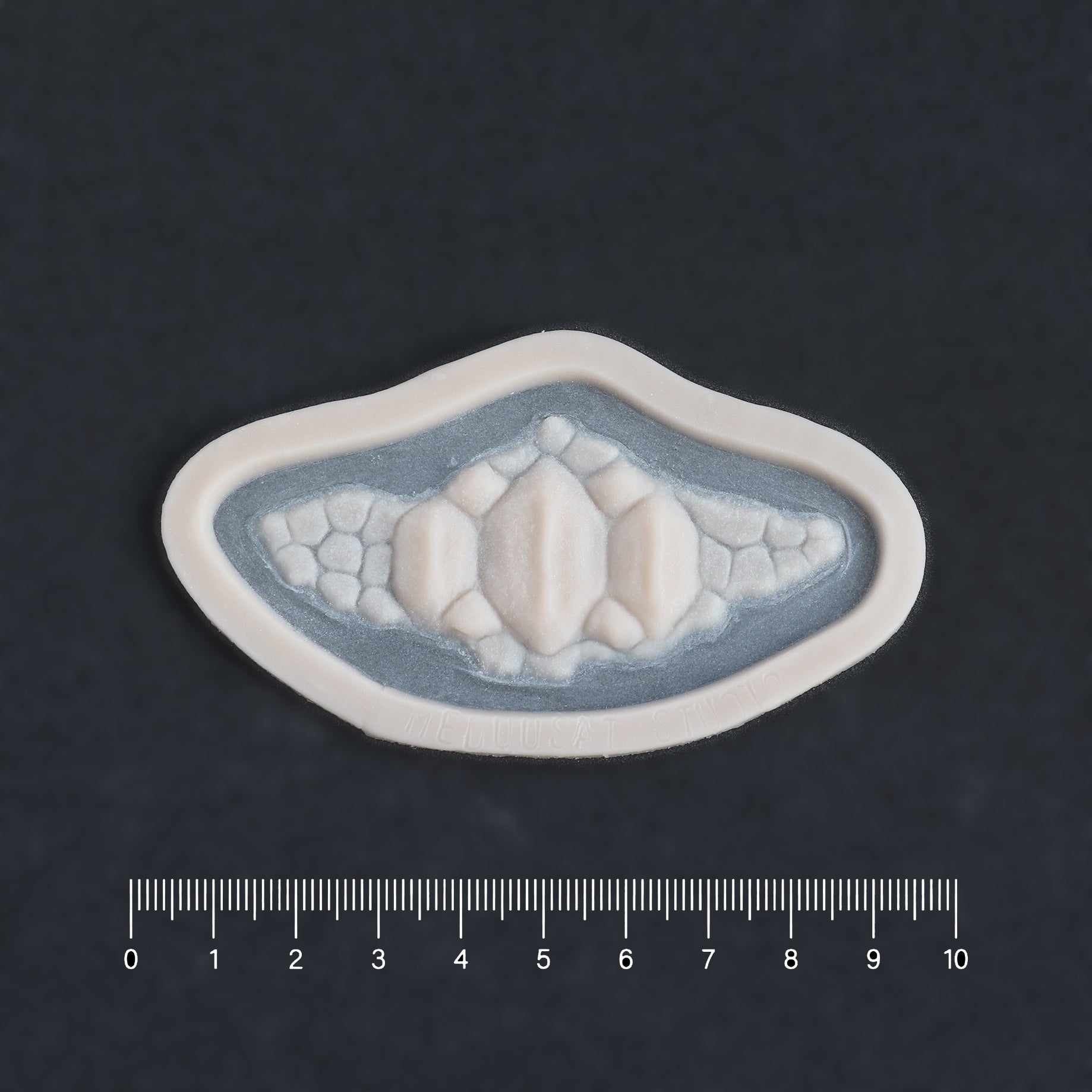 Alligator Scales chin piece in vanilla shade on a black surface with a ruler.